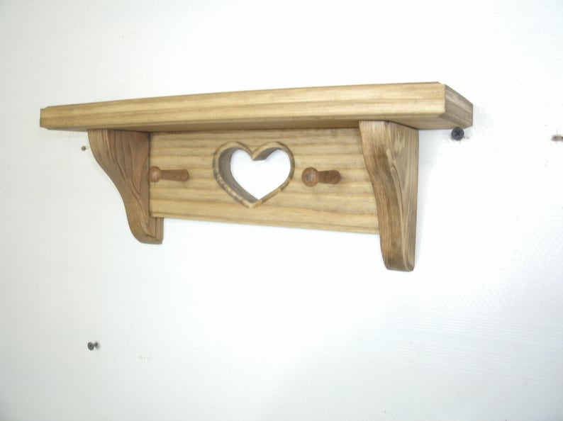 May include: A wooden shelf with a heart-shaped cutout and two pegs below. The shelf is made of light-colored wood with a natural finish. The shelf is mounted on a white wall.