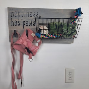 Puede incluir: Un letrero de madera con el texto "Happiness has paws" colgado en una pared. El letrero tiene dos ganchos para colgar correas y una cesta de alambre para guardar juguetes para perros. La cesta contiene una pelota de b&eacute;isbol, un juguete de peluche verde y un juguete de cuerda multicolor.
