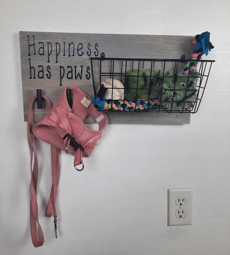 Puede incluir: Un letrero de madera con el texto "Happiness has paws" colgado en una pared. El letrero tiene dos ganchos para colgar correas y una cesta de alambre para guardar juguetes para perros. La cesta contiene una pelota de b&eacute;isbol, un juguete de peluche verde y un juguete de cuerda multicolor.