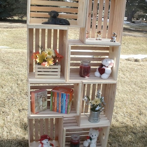 Op de afbeelding: Een houten krattenrek met meerdere planken, elk met verschillende items, waaronder een zwart hondenbeeldje, een boeket bloemen, een boekensets, een teddybeer en een kleine gieter met bloemen.