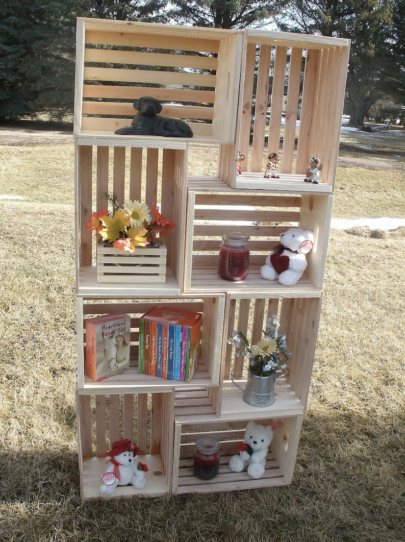 Op de afbeelding: Een houten krattenrek met meerdere planken, elk met verschillende items, waaronder een zwart hondenbeeldje, een boeket bloemen, een boekensets, een teddybeer en een kleine gieter met bloemen.