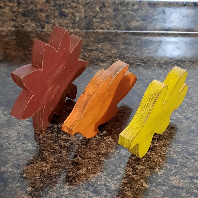 May include: Three wooden leaf-shaped decorations in shades of red, orange, and yellow. The rustic, distressed finish gives them a vintage look. These decorative leaves are likely for fall or autumn-themed decor.