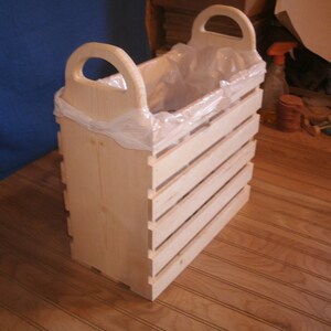 May include: A wooden trash can with a plastic liner and two handles. The can is made of natural wood and has a slatted design.