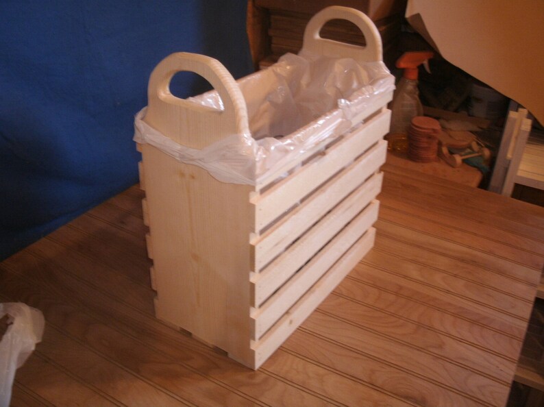 May include: A wooden trash can with a plastic liner and two handles. The can is made of natural wood and has a slatted design.