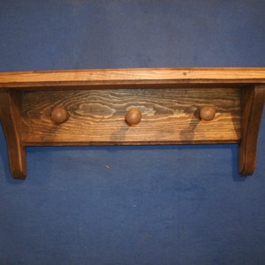 May include: A wooden shelf with three hooks, stained in a dark brown color. The shelf has a simple design with rounded edges and a slight curve on the sides.