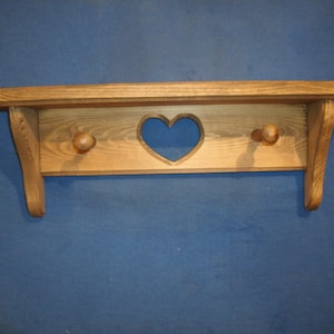 May include: A wooden shelf with a heart-shaped cutout and two wooden knobs. The shelf is made of light-colored wood and has a rustic, handcrafted appearance. The shelf is designed for wall mounting.