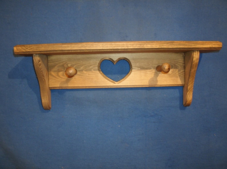 May include: A wooden shelf with a heart-shaped cutout and two wooden knobs. The shelf is made of light-colored wood and has a rustic, handcrafted appearance. The shelf is designed for wall mounting.