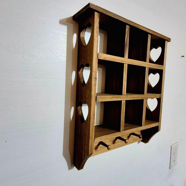 May include: A wooden wall shelf with a heart-shaped cutout design. The shelf has three tiers and three hooks on the bottom. The shelf is stained a dark brown color.