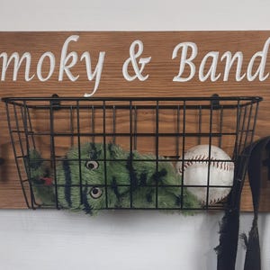 Puede incluir: Un letrero de madera con el texto "Smoky & Bandit" en letras blancas. Una cesta de alambre negro est&aacute; unida al letrero y contiene un animal de peluche verde y negro y una pelota de b&eacute;isbol blanca. Dos ganchos negros est&aacute;n unidos al letrero.