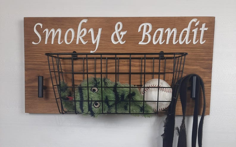 Puede incluir: Un letrero de madera con el texto "Smoky & Bandit" en letras blancas. Una cesta de alambre negro est&aacute; unida al letrero y contiene un animal de peluche verde y negro y una pelota de b&eacute;isbol blanca. Dos ganchos negros est&aacute;n unidos al letrero.