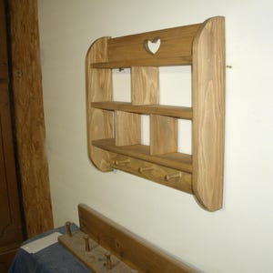 K&ouml;nnte beinhalten: Wandregal aus Holz mit drei Regalen und vier Haken darunter. Das Regal hat einen herzf&ouml;rmigen Ausschnitt oben. Das Holz hat eine nat&uuml;rliche Oberfl&auml;che. Das Regal ist an einer wei&szlig;en Wand befestigt.