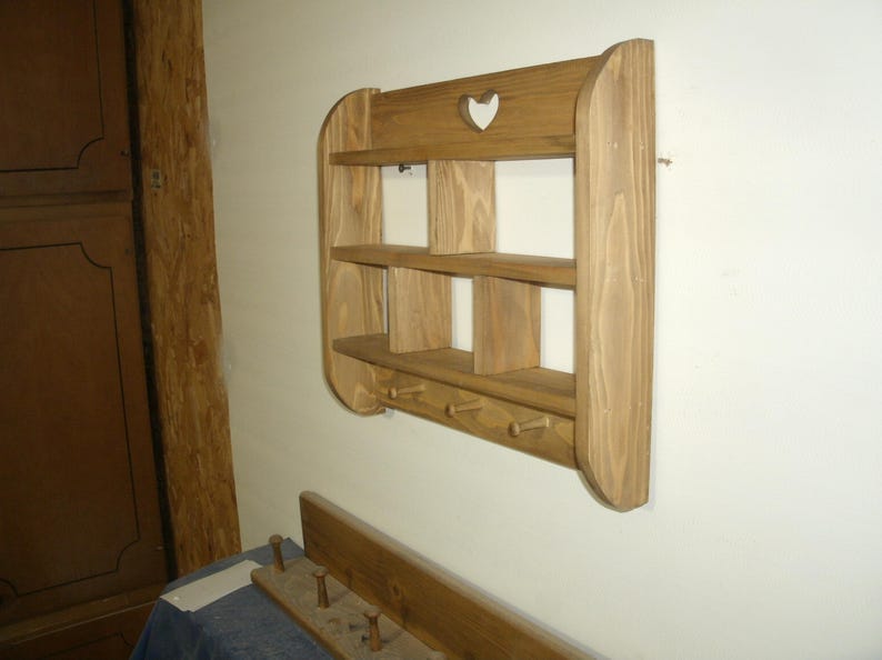 K&ouml;nnte beinhalten: Wandregal aus Holz mit drei Regalen und vier Haken darunter. Das Regal hat einen herzf&ouml;rmigen Ausschnitt oben. Das Holz hat eine nat&uuml;rliche Oberfl&auml;che. Das Regal ist an einer wei&szlig;en Wand befestigt.