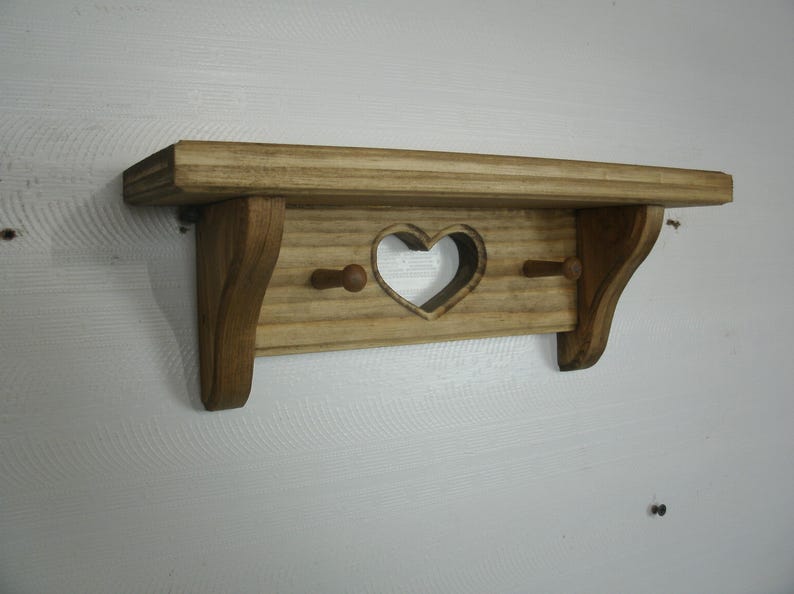 May include: Wooden shelf with a heart-shaped cutout and two pegs. The shelf is made of light-colored wood and has a rustic, country style. The shelf is approximately 12 inches wide.