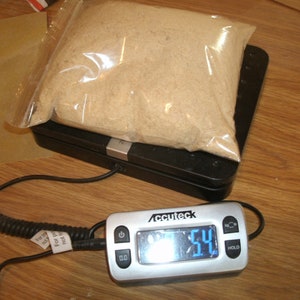 Pode incluir: Uma balan&ccedil;a de cozinha digital com um visor branco e prateado mostrando um peso de 54 g. Um saco de pl&aacute;stico transparente contendo um p&oacute; bege est&aacute; sobre a balan&ccedil;a.