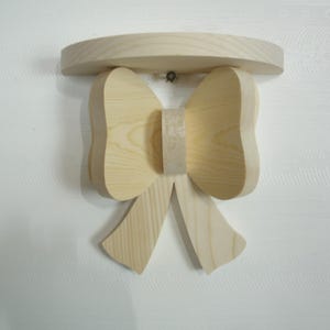 May include: A wooden wall shelf in the shape of a bow. The shelf is made of unfinished light-colored wood and has a rounded top. The bow design is centered below the shelf, with two loops and two hanging tails.