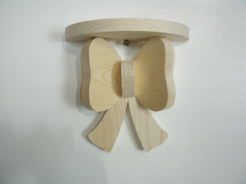 May include: A wooden wall shelf in the shape of a bow. The shelf is made of unfinished light-colored wood and has a rounded top. The bow design is centered below the shelf, with two loops and two hanging tails.