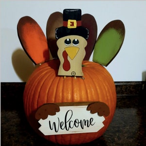 May include: A wooden turkey figurine with a black hat and a welcome sign, perched on top of an orange pumpkin.