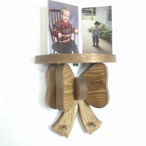May include: Wooden wall shelf with a bow-shaped design. The shelf holds two framed photos of children. The bow is made of layered wood with a natural finish, and the shelf is a circular shape.