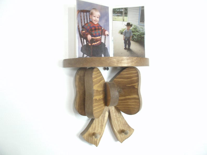 May include: Wooden wall shelf with a bow-shaped design. The shelf holds two framed photos of children. The bow is made of layered wood with a natural finish, and the shelf is a circular shape.