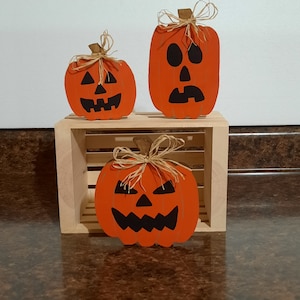 May include: Three orange wooden jack-o'-lanterns with black facial features and raffia accents. The pumpkins are displayed on a small wooden crate. The pumpkins have different expressions, perfect for Halloween decor.