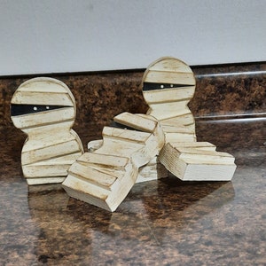 May include: Three wooden mummy figurines with black eyes and white painted bandages. The mummies are arranged in a pile on a brown surface.