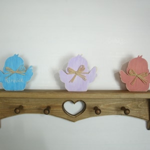 May include: A wooden shelf with a heart-shaped cutout in the front. Three wooden bird figurines are displayed on the shelf. The birds are painted in shades of blue, purple, and pink. The blue bird has the name "Kinrick" written on it.