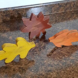 May include: Three wooden leaf cutouts in autumn colors: yellow, orange, and brown. The leaf shapes are stylized and appear to be hand-painted, suggesting a craft or decorative item. The leaves are arranged on a brown surface.