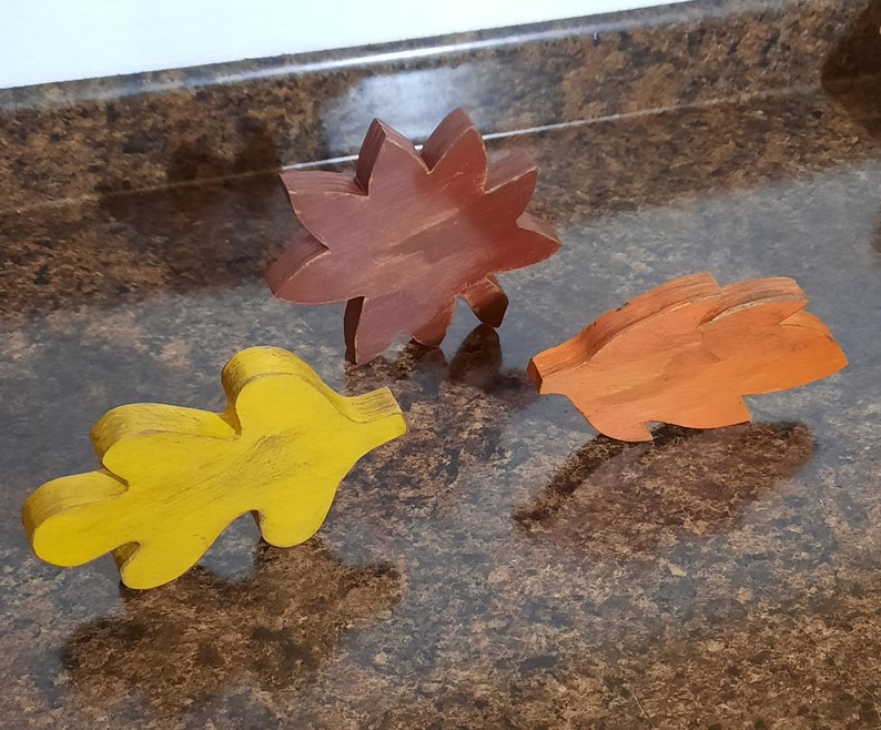 May include: Three wooden leaf cutouts in autumn colors: yellow, orange, and brown. The leaf shapes are stylized and appear to be hand-painted, suggesting a craft or decorative item. The leaves are arranged on a brown surface.