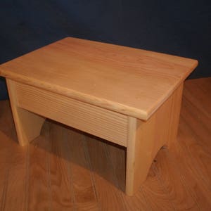 May include: A wooden stool with a natural finish. The stool has a simple design with a rectangular top and four legs.