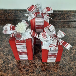 May include: Three red and white striped wooden boxes decorated with white and red ribbon and white pine cone ornaments. The boxes are stacked on top of each other.