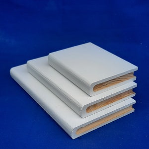 May include: Three wooden book-shaped objects, painted white, are stacked on a blue background. The objects vary in size, with the largest at the bottom and the smallest at the top. The edges are rounded, and the inside reveals natural wood.