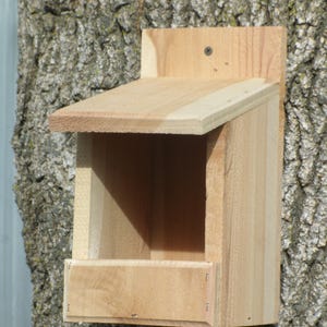 May include: A wooden birdhouse with a slanted roof and a small opening for birds to enter. The birdhouse is attached to a tree trunk.