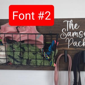 Puede incluir: Un organizador de pared de madera con el texto "The Samsel Pack" en fuente cursiva blanca. El organizador tiene una cesta de alambre negra con un collar de perro rosa, un juguete de peluche verde y una pelota de b&eacute;isbol. Hay dos ganchos negros con correas rosas y negras colgando.