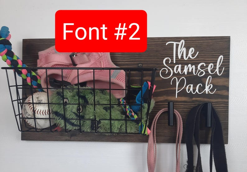 Puede incluir: Un organizador de pared de madera con el texto "The Samsel Pack" en fuente cursiva blanca. El organizador tiene una cesta de alambre negra con un collar de perro rosa, un juguete de peluche verde y una pelota de b&eacute;isbol. Hay dos ganchos negros con correas rosas y negras colgando.