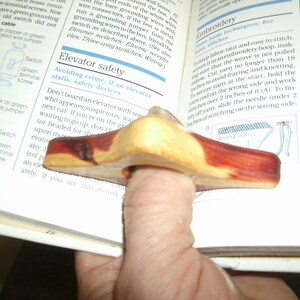 Puede incluir: Un sujetalibros de madera en forma de anillo para el pulgar, con una mancha marr&oacute;n rojiza. El anillo est&aacute; dise&ntilde;ado para ajustarse a un dedo y mantener un libro abierto.