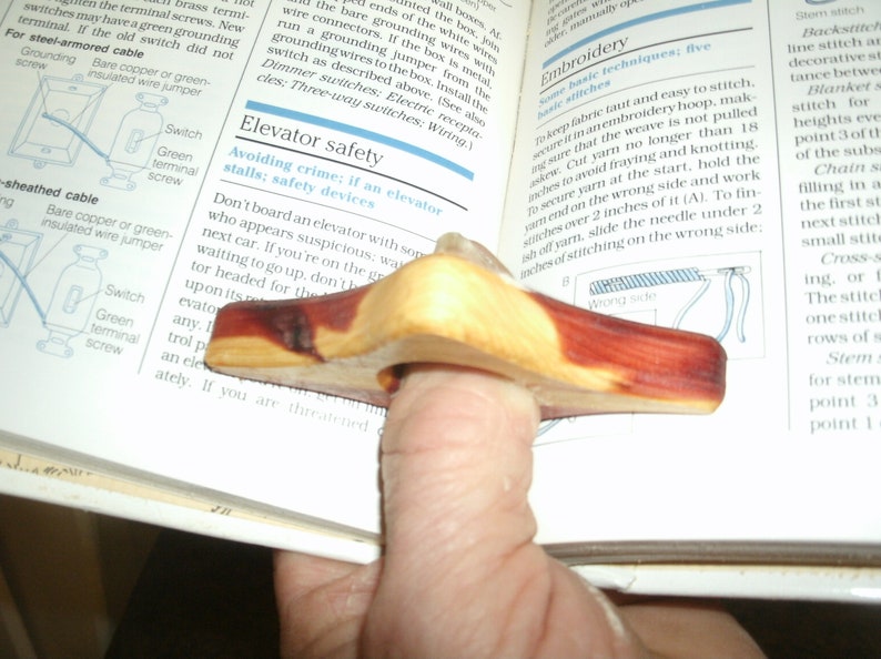 Puede incluir: Un sujetalibros de madera en forma de anillo para el pulgar, con una mancha marr&oacute;n rojiza. El anillo est&aacute; dise&ntilde;ado para ajustarse a un dedo y mantener un libro abierto.