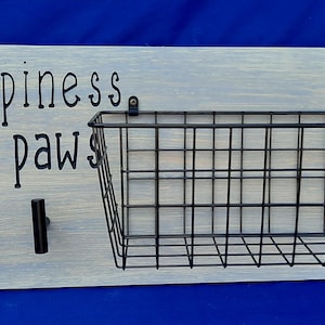 Puede incluir: Un letrero de madera blanco con el texto "Happiness has paws" y dos ganchos negros. Una cesta de alambre negro est&aacute; unida al letrero.