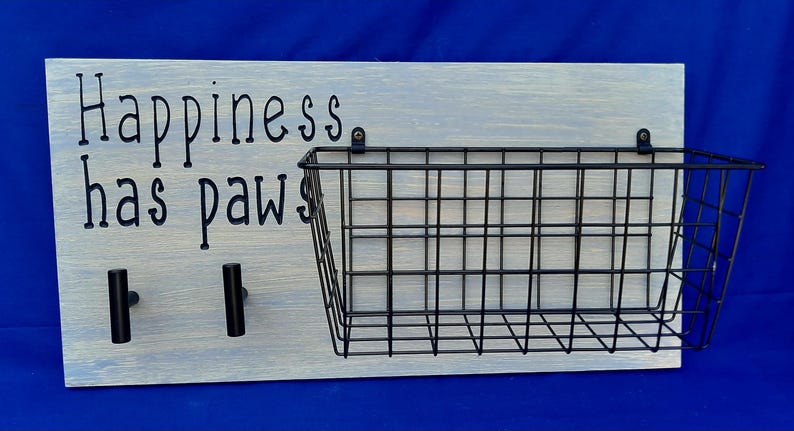 Puede incluir: Un letrero de madera blanco con el texto "Happiness has paws" y dos ganchos negros. Una cesta de alambre negro est&aacute; unida al letrero.