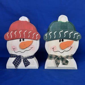 Op de afbeelding: Twee houten sneeuwpopfiguren met geschilderde gezichten, rode en groene hoeden en geruite sjaals. De sneeuwpoppen staan op een blauwe achtergrond.