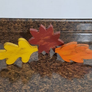 May include: Three wooden fall leaf decorations in yellow, orange, and red. The yellow leaf is a simple oak leaf shape. The orange leaf has a maple leaf shape. The red leaf has a star shape. These are fall-themed decorations.
