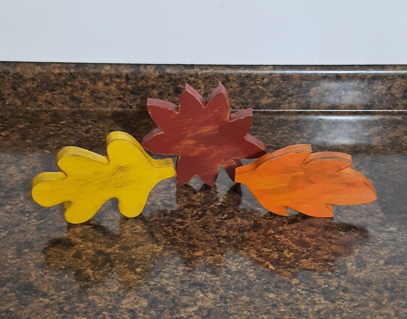 May include: Three wooden fall leaf decorations in yellow, orange, and red. The yellow leaf is a simple oak leaf shape. The orange leaf has a maple leaf shape. The red leaf has a star shape. These are fall-themed decorations.