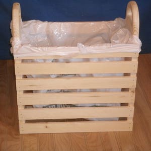 May include: A wooden crate with a white plastic bag inside. The crate has two handles on the top and is made of natural wood.
