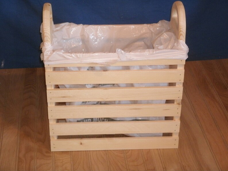 May include: A wooden crate with a white plastic bag inside. The crate has two handles on the top and is made of natural wood.
