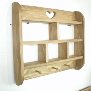 K&ouml;nnte beinhalten: Wandregal aus Holz mit nat&uuml;rlichem Finish. Das Regal hat drei Ebenen, F&auml;cher und drei Haken. Oben befindet sich ein herzf&ouml;rmiger Ausschnitt. Dieses dekorative Regal ist perfekt f&uuml;r die Pr&auml;sentation kleiner Gegenst&auml;nde.