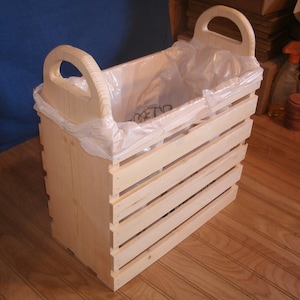 May include: A wooden trash can with two handles and a plastic trash bag inside. The can is made of natural wood and has a slatted design.