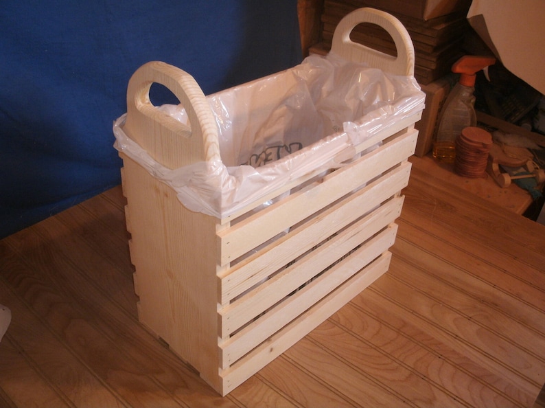 May include: A wooden trash can with two handles and a plastic trash bag inside. The can is made of natural wood and has a slatted design.