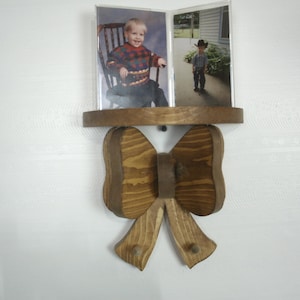 May include: Wooden photo display with a shelf and a bow-tie design. The shelf holds two photos of a young child. The bow-tie has two pegs for hanging items. The wood has a natural finish.