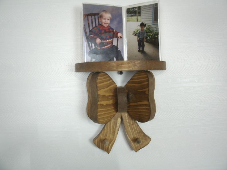 May include: Wooden photo display with a shelf and a bow-tie design. The shelf holds two photos of a young child. The bow-tie has two pegs for hanging items. The wood has a natural finish.