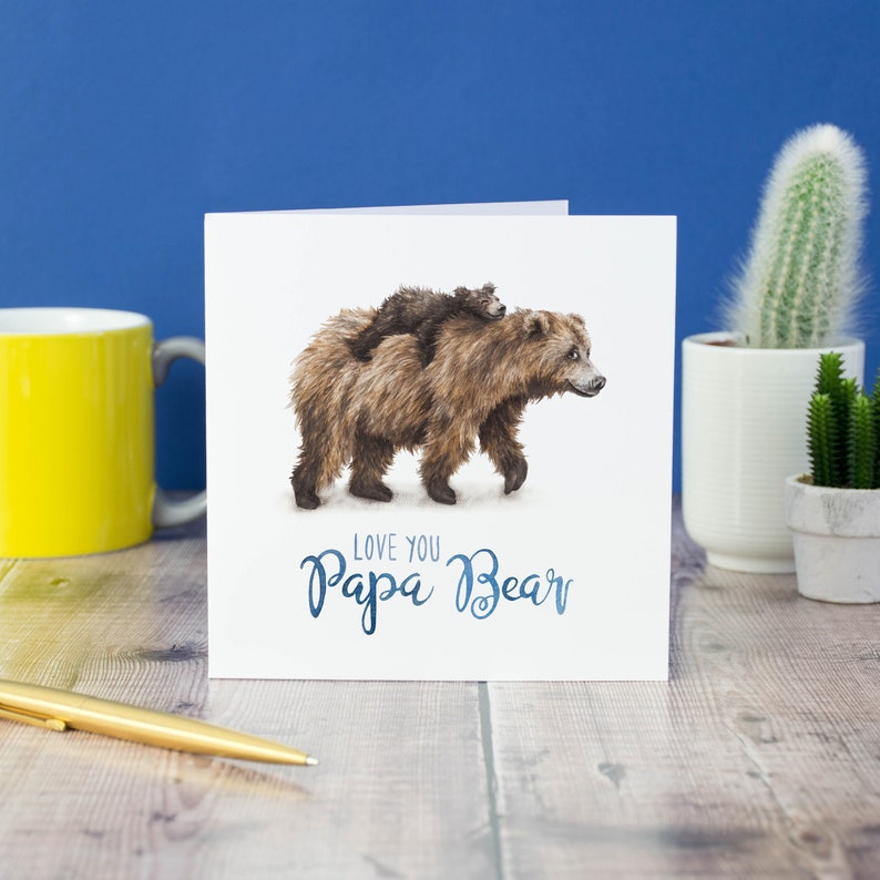 Peut inclure: Une carte blanche avec un ours brun et un petit ourson brun sur son dos. Le texte "Love you Papa Bear" est &eacute;crit en lettres cursives bleues.