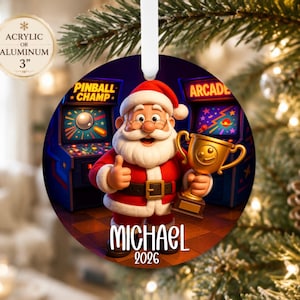 May include: A round 3-inch acrylic or aluminum Christmas ornament featuring a cartoon Santa Claus holding a gold trophy in an arcade. Santa wears a red suit with a black belt and gives a thumbs up. The text reads PINBALL CHAMP, ARCADE, MICHAEL, 2026.
